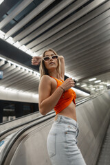 Wall Mural - Cool trendy stylish pretty woman hipster with fashion white sunglasses in trendy casual clothes with orange top and jeans on the escalator in the subway