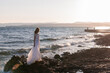 © Iuliia Pilipeichenko - Beautiful young bride in a white wedding dress stands on the ocean or sea in the sun. Fashion shooting in a wedding or evening dress