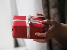 Close Up Side View Of Woman Hands Out Red Gift Box With White Silk Ribbon, Giving Present On Holiday. Merry Christmas, Happy New Year, Valentine's Day, Holiday, Festive, Birthday, Surprises Concept.