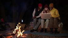 Cheerful Multiracial Friends Using Laptop Together At Camping In The Forest. Three People Sitting Near A Bonfire Enjoying The Night, Surfing In The Net, Watching A Movie. Cozy Copy Space Image.