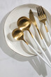 Matte gold flatware set with white handles, fork, spoon, knife and teaspoon on ceramic plate, top view