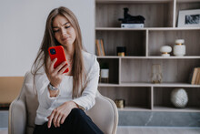 Frustrated Caucasian Blonde Young Woman Sitting On Chair Looks At Phone  With Unhappy Face Expression Reads Message At Home. Beautiful Swedish Businesswoman Disapointed After Video Call. Failure