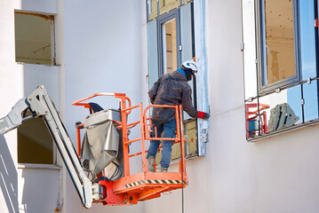 Wall Mural - Man in lift bucket caulk, seal exterior window with putty knife, maintain exterior window. Worker in lifting platform at height insulate and seal around building windows. Worker in construction cradle