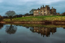 Scenic View Of Ripley Castle Reflecting In A Lake Against A Cloudy Sky In England.