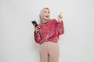 Excited Asian woman wearing pink sweater and hijab pointing at the copy space on top of her while holding her phone, isolated by white background