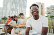 © Jose Calsina - On foreground a smiling cheerful young African american student with a positive smile, white perfect teeth, looking at camera sitting outside taking a break at the university. at the background a