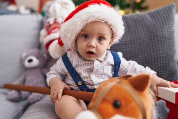 Sticker - Adorable caucasian baby holding horse toy sitting on sofa by christmas tree at home