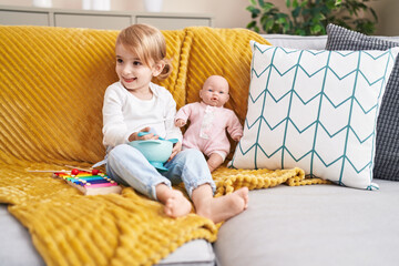 Sticker - Adorable caucasian girl playing with baby doll sitting on sofa at home