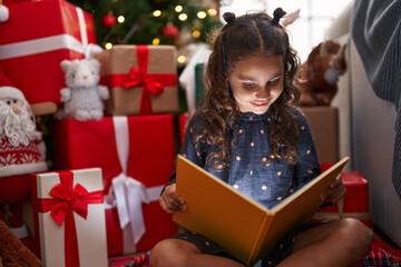 Sticker - Adorable hispanic girl reading book sitting on floor by christmas tree at home