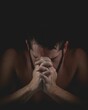 Shirtless male with beard kneeling with hands folded and head bowed to pray with black background
