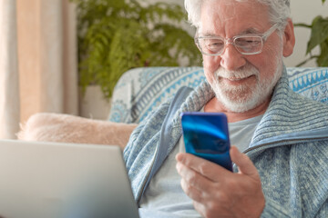 Wall Mural - Smiling elderly bearded man sitting on sofa at home with laptop while talking on mobile phone. Senior in eyeglasses and light blue sweater