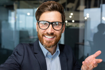 Wall Mural - Businessman looks into smartphone camera and talks on video call, man uses phone for remote communication with friends and colleagues, investor boss in glasses works inside office.