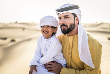 Wall Mural - Playful arab man and his son wering traditional middle eastern emirate clothing playing and having fun in the desert of Dubai