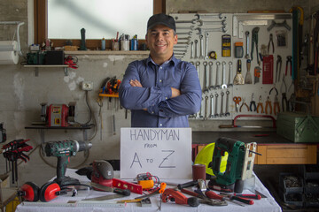 Canvas Print - Image of a smiling man in his workshop behind a bench full of work tools with handyman from A to Z written on it. Man who can fix and fix everything
