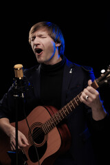 Male musician with guitar in hands playing and posing on black background in blue scenic light