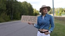 Young Woman Actively Look Out Passing Cars With Cardboard Poster On Roadside Of Empty Road. Lady In Hat And Denim Outfit Escape From City To Go Anywhere. Travelling, Freedom, Hitchhiking, Vacations
