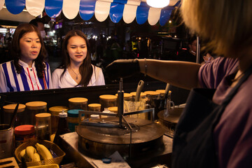 Selective focus of crepe shop and asian young women friends choose order crape cake and wait cooking dessert on hot pan at downtown street food city, delicious food serve for tourist or night travel.