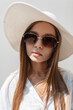 © alones - Fashionable summer female portrait of a beautiful woman with sunglasses in a fashion hat and white elegant shirt with sunlight on the street