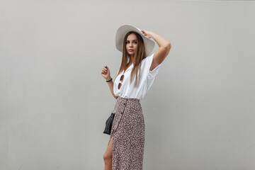 Fashionable beautiful stylish elegant woman with a trendy straw hat in a fashion white shirt with a skirt and a handbag near a gray wall in the city