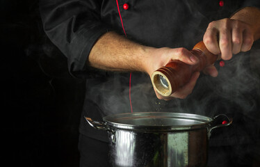 The chef adds ground pepper to a pot of boiling food. Retsoran kitchen cooking concept with advertising space on black background.