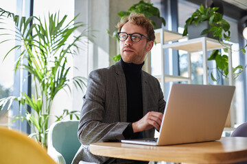 Portrait of young attractive businessman wearing stylish eyeglasses and suit using laptop computer working online sitting at workplace. Successful business