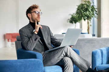 Pensive businessman working using laptop planning startup  sitting in modern office. Portrait of young confident freelancer sitting at workplace 