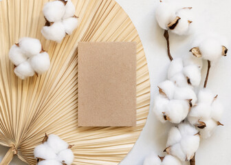 Wall Mural - Blank kraft paper card on dried palm leaf with cotton flowers top view, wedding mockup