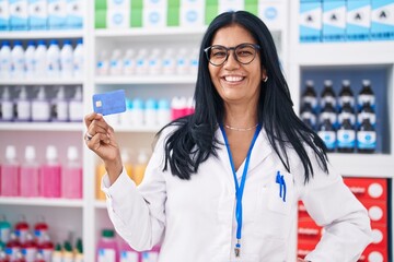 Sticker - Mature hispanic woman working at pharmacy drugstore holding credit card looking positive and happy standing and smiling with a confident smile showing teeth