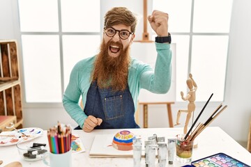 Sticker - Redhead man with long beard painting clay bowl at art studio angry and mad raising fist frustrated and furious while shouting with anger. rage and aggressive concept.