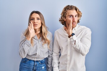 Poster - Young couple standing over blue background hand on mouth telling secret rumor, whispering malicious talk conversation