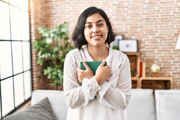 Wall Mural - Young latin woman smiling confident hugging book at home