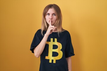 Sticker - Blonde caucasian woman wearing bitcoin t shirt asking to be quiet with finger on lips. silence and secret concept.