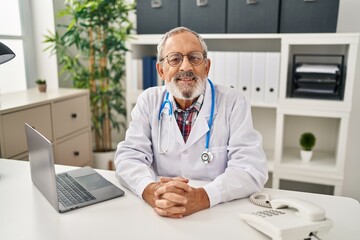 Wall Mural - Senior grey-haired man doctor smiling confident sitting on table at clinic