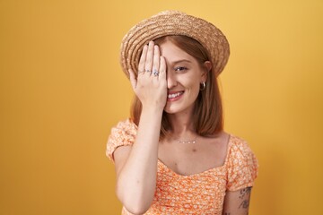 Poster - Young redhead woman standing over yellow background wearing summer hat covering one eye with hand, confident smile on face and surprise emotion.