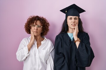 Canvas Print - Hispanic mother and daughter wearing graduation cap and ceremony robe touching mouth with hand with painful expression because of toothache or dental illness on teeth. dentist