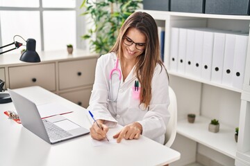 Canvas Print - Young beautiful hispanic woman doctor using laptop writing on covid certificate card at clinic