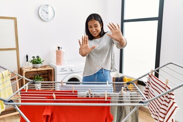 Poster - Young hispanic woman putting fresh laundry on clothesline afraid and terrified with fear expression stop gesture with hands, shouting in shock. panic concept.