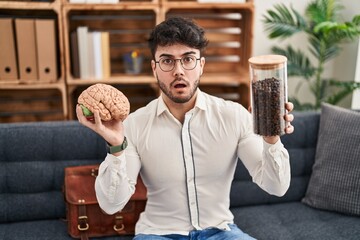 Sticker - Hispanic man with beard working at therapy office holding brain and coffee beans in shock face, looking skeptical and sarcastic, surprised with open mouth