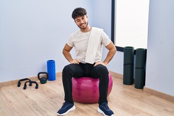 Sticker - Hispanic man with beard sitting on pilate balls at yoga room winking looking at the camera with sexy expression, cheerful and happy face.