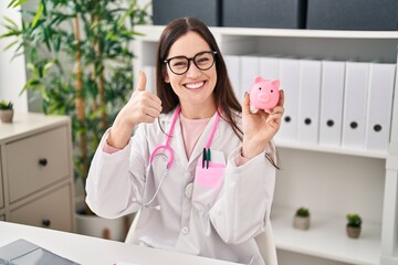 Poster - Young doctor woman holding piggy bank smiling happy and positive, thumb up doing excellent and approval sign