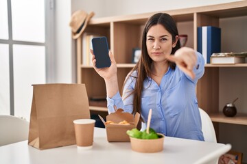 Poster - Young brunette woman eating take away food at home showing smartphone screen pointing with finger to the camera and to you, confident gesture looking serious