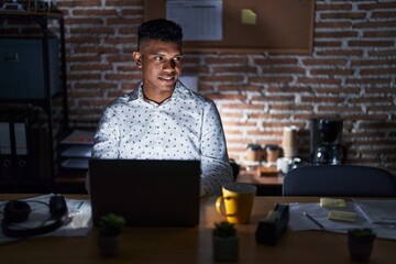 Poster - Young hispanic man working at the office at night looking away to side with smile on face, natural expression. laughing confident.