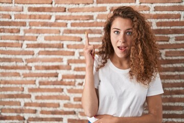 Poster - Young caucasian woman standing over bricks wall background pointing finger up with successful idea. exited and happy. number one.