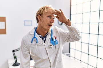 Canvas Print - Young blond man wearing doctor uniform and stethoscope at clinic very happy and smiling looking far away with hand over head. searching concept.