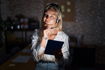 Wall Mural - Young blonde woman working at the office at night with hand on chin thinking about question, pensive expression. smiling and thoughtful face. doubt concept.