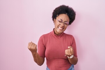 Wall Mural - Beautiful african woman with curly hair standing over pink background very happy and excited doing winner gesture with arms raised, smiling and screaming for success. celebration concept.