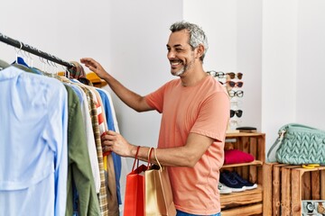 Middle age grey-haired man customer smiling confident holding clothes of rack at clothing store