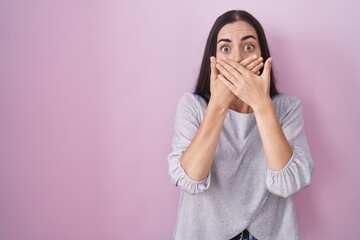 Poster - Young brunette woman standing over pink background shocked covering mouth with hands for mistake. secret concept.