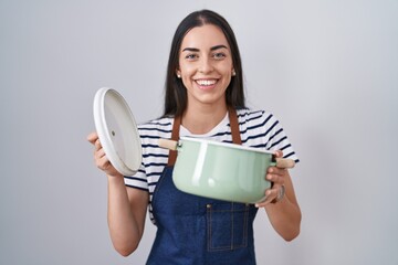 Poster - Young brunette woman wearing apron holding cooking pot smiling with a happy and cool smile on face. showing teeth.