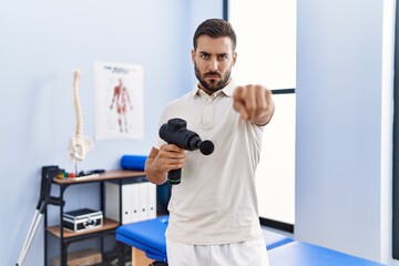 Poster - Handsome hispanic man holding therapy massage gun at physiotherapy center pointing with finger to the camera and to you, confident gesture looking serious